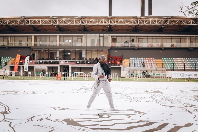 A man dressed in all white stands in the middle of a large drawing in black permeant marker in the middle of a football field.