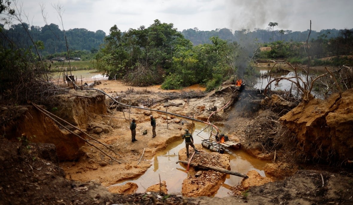 brazil-gold-munduruku A muddy area is shown, with three people standing in the middle of it. Trees are visible past them, with a river behind it.