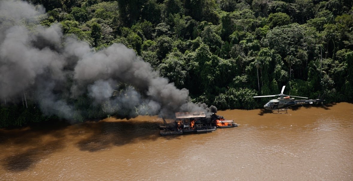 brazil-gold Grey smoke billows out from a boat on a wide brown river. A helicopter hovers a short distance away.
