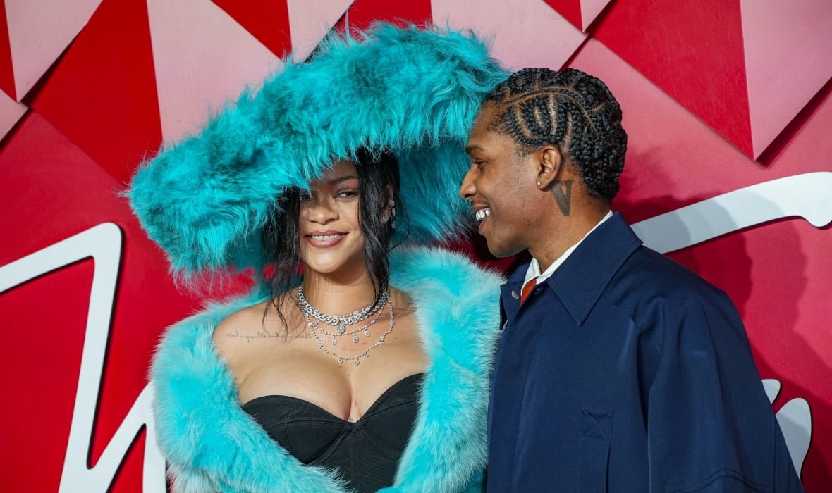 britain-the-fashion-awards A couple are shown on a red carpet, posing in front of a red backdrop. The woman is wearing a massive, cyan feathered hat that matches the bright cyan fluffy coat she is wearing over a black dress. She is smiling at the camera, while the man smiles at her.