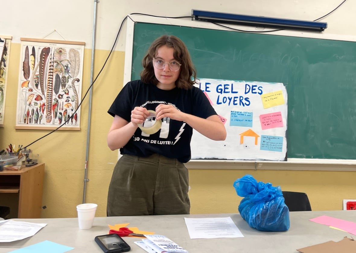 catherine-fournier A young woman with glasses stands at a table holding a roll of tape in front of a chalk board.