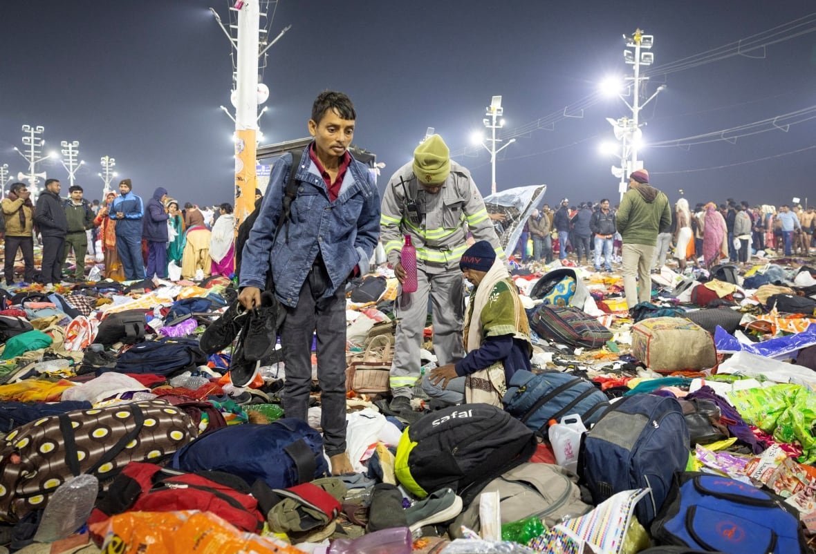 india-religion-kumbh-stampede A man in a blue denim jacket stands amid a pile of clothing and bags on the ground. Behind him, an older man kneels on the ground.