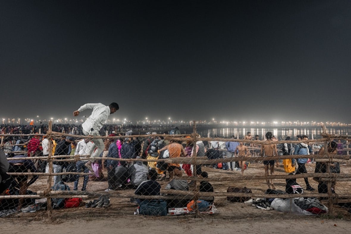india-stampede A man dressed in white jumped over a chain link fence on the bank of a river. Dozens of people are on the river bank.
