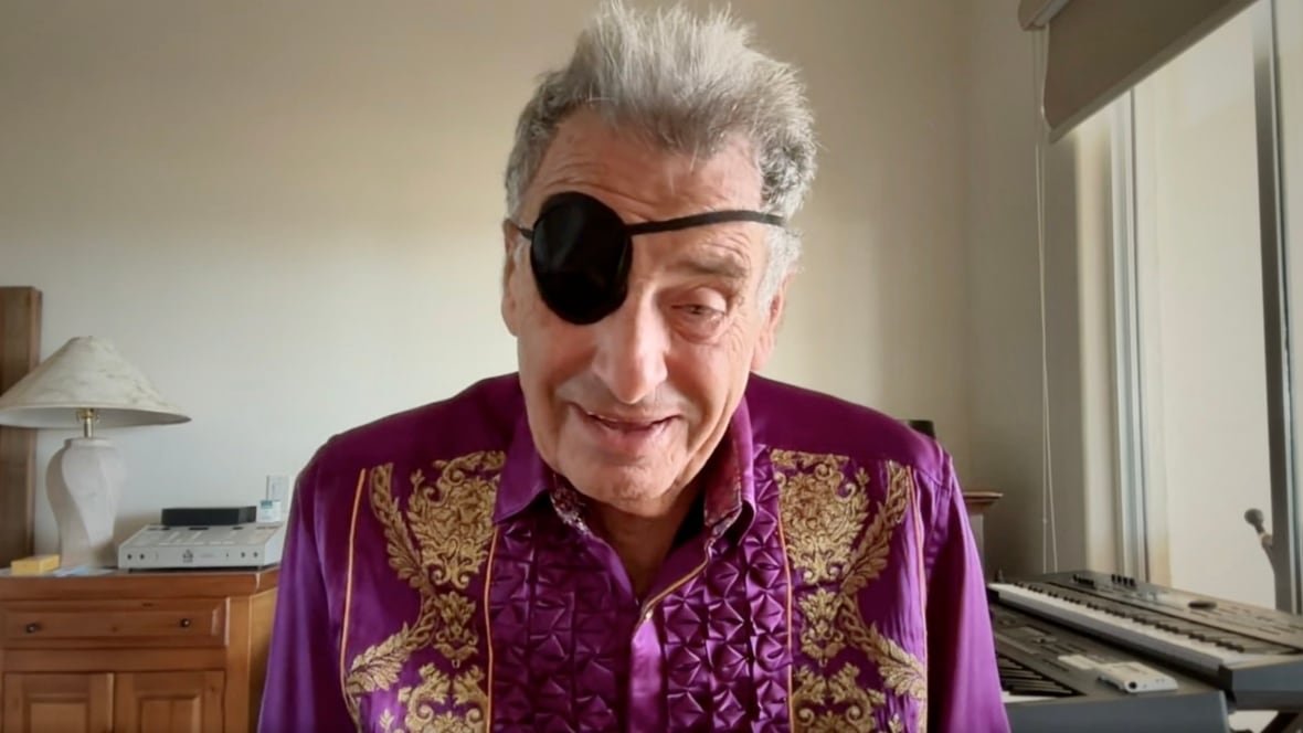 jack-herschorn A man in a purple silk collared shirt with an eye patch and grey hair looks at the camera from a bedroom.