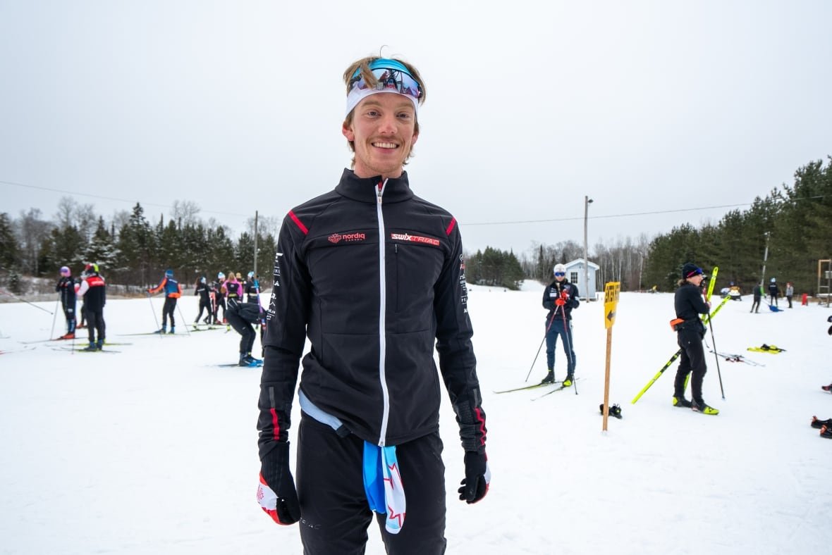 max-hollmann A man wearing winter clothing smiles as he poses for a photo outdoors, with cross-country skiers behind him.