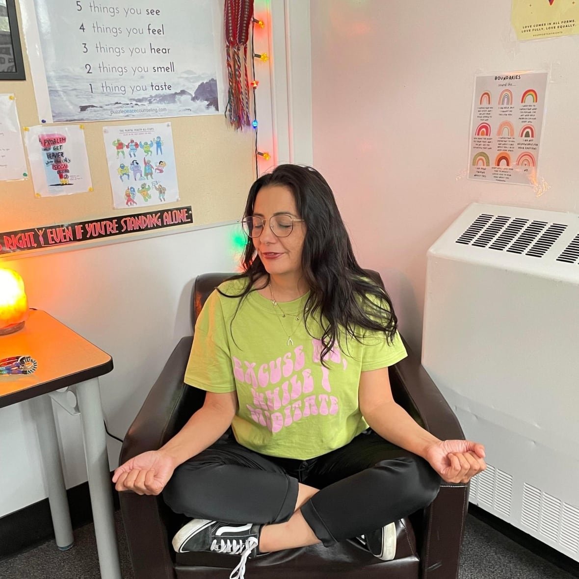 nikki-houde-1 A woman sitting on a leather chair, with her legs crossed, her arms on her knees and her hands turned upwards. She looks calm and collected, sitting in the lotus position.