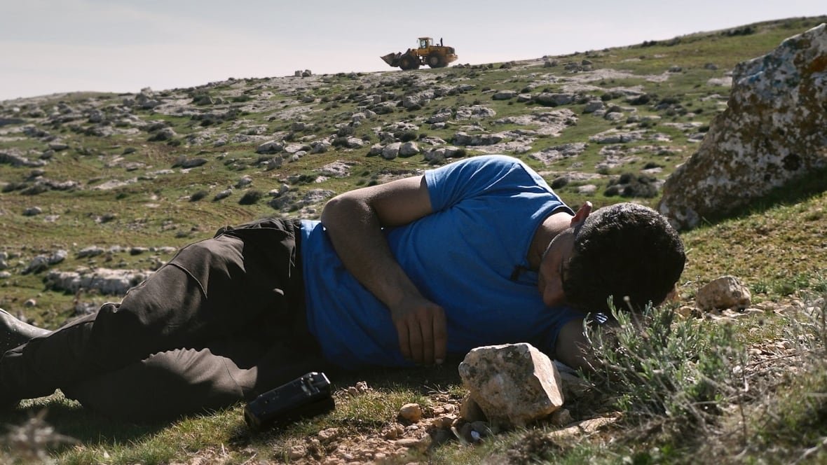 no-other-land-1 A man lies on the ground on a grassy hill studded with rocks. A bulldozer is visible at the top of the hill beyond him.