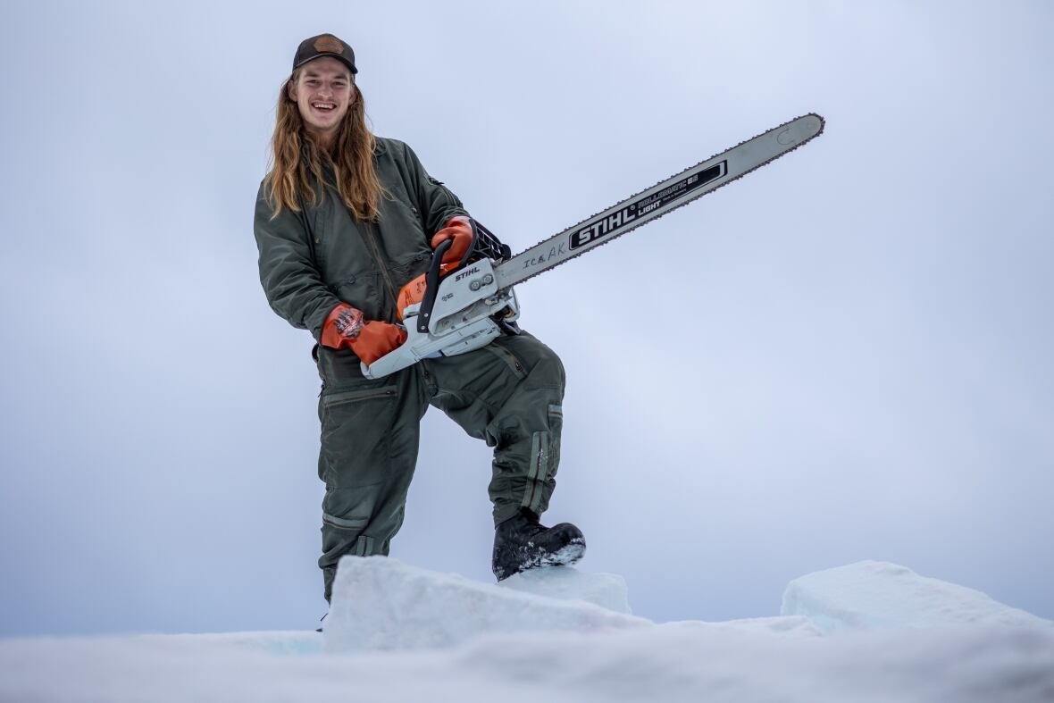 riley-knaus A man with long hair and heavy winter attire smiling while holding a chainsaw.