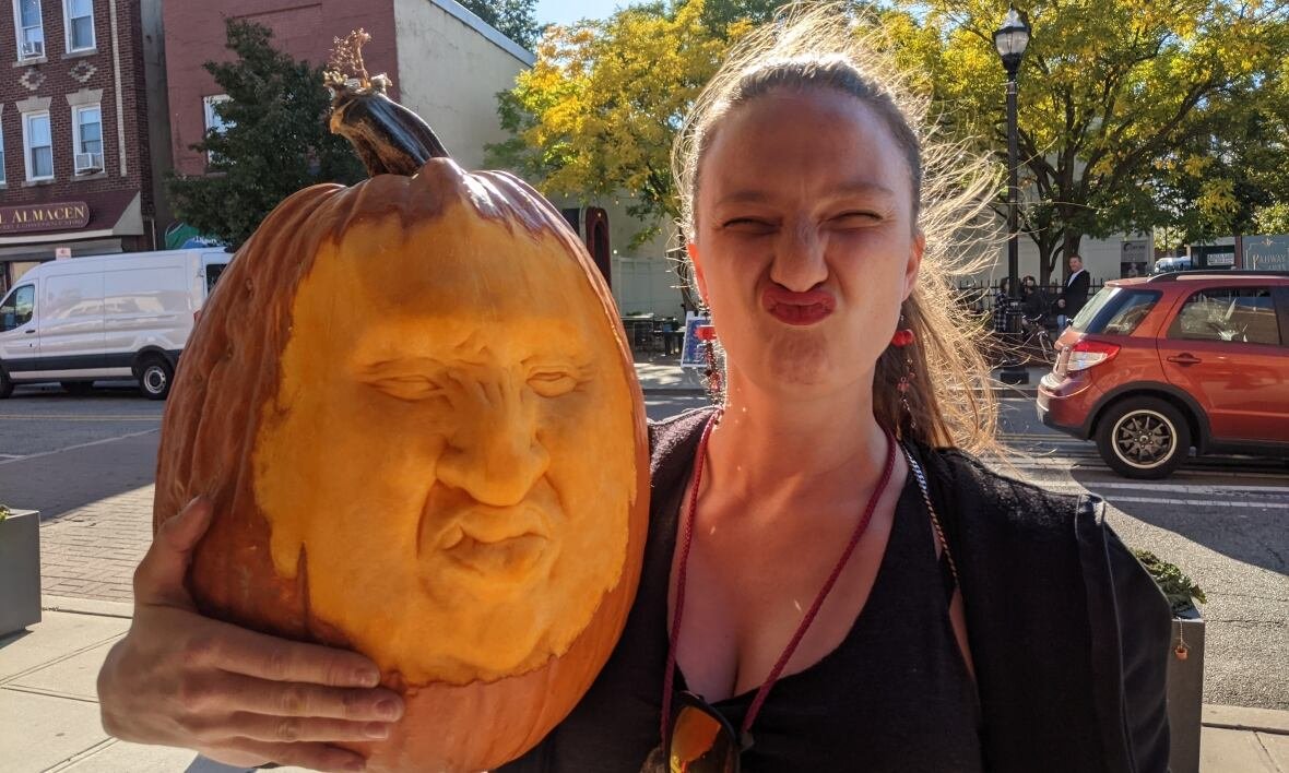 sarah-blostein-pumpkin A woman making a wrinkly face while holding a pumpkin with a similar face expression carved into it.