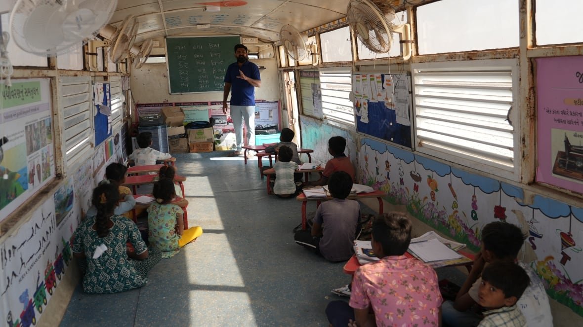 A teacher stands in front of a blackboard talking to students seated on the floor inside a bus.