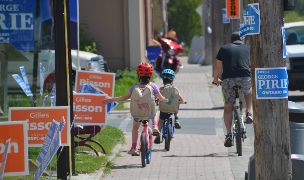 timmins-provincial-election-2022 Two kids and an adult bike down a sidewalk lined with election signs