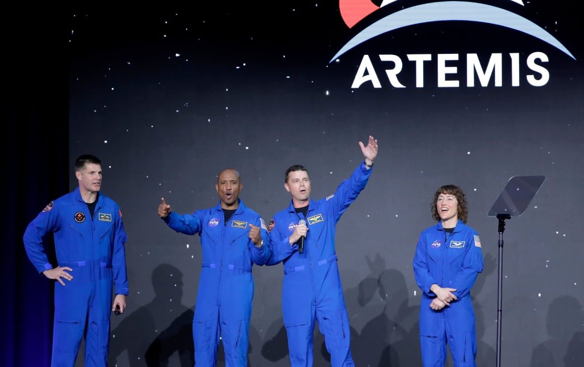Four astronauts in blue outfits stand together, some mid-whoop, others smiling. An Artemis logo is visible on the wall behind them.