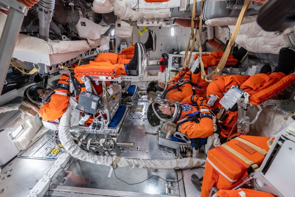 Four people in orange spacesuits are seen lying in a spacecraft.