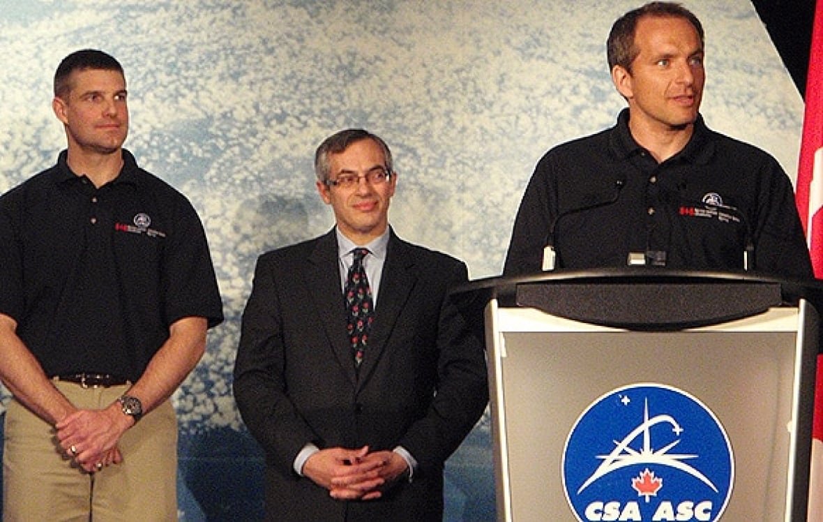 Three men are seen on a stage, with one standing at a lecturn.