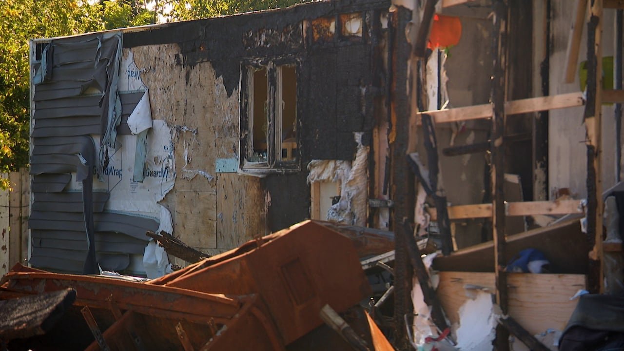 Melted siding exposes wooden walls on the side of a small home. There is charred materials around the side of the home.