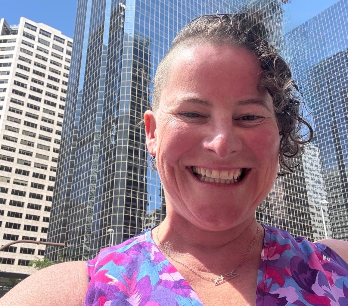 default-494 A woman with short curly hair shaved on one side smiles broadly with tall office buildings in the background.