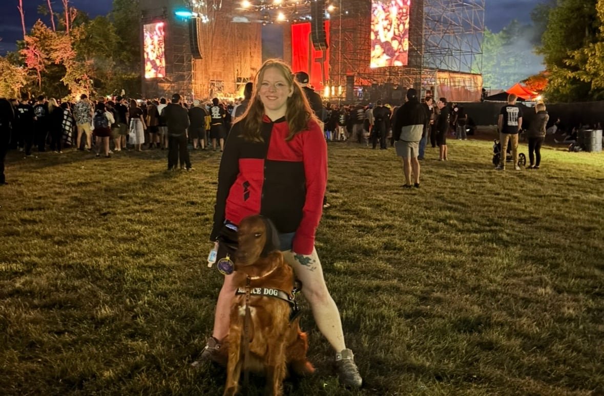 default-132 A woman stands in the crowd at an outdoor concert, her golden retriever standing between her legs.