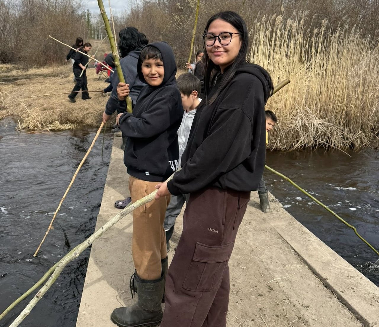 default-457 Cheerful children stand on a wooden bridge over a stream, holding lengthy sticks.