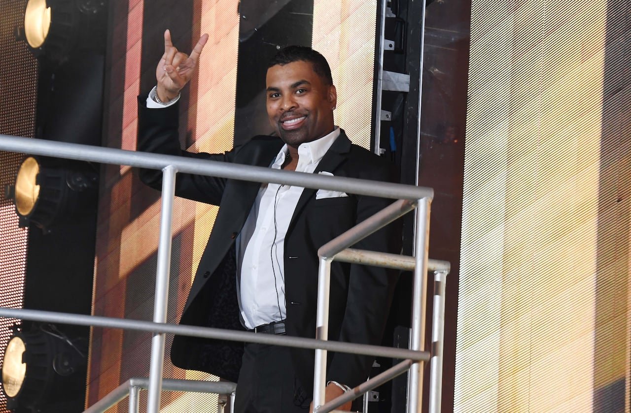 default-304 A smiling man in a dark suit gives a "rock on" gesture, extending index and pinky fingers while curling his thumb over bent middle and ring fingers.
